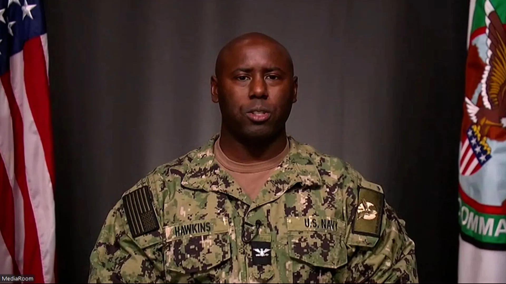 U.S. Navy servicemember in camouflage speaking during a televised briefing between two flags