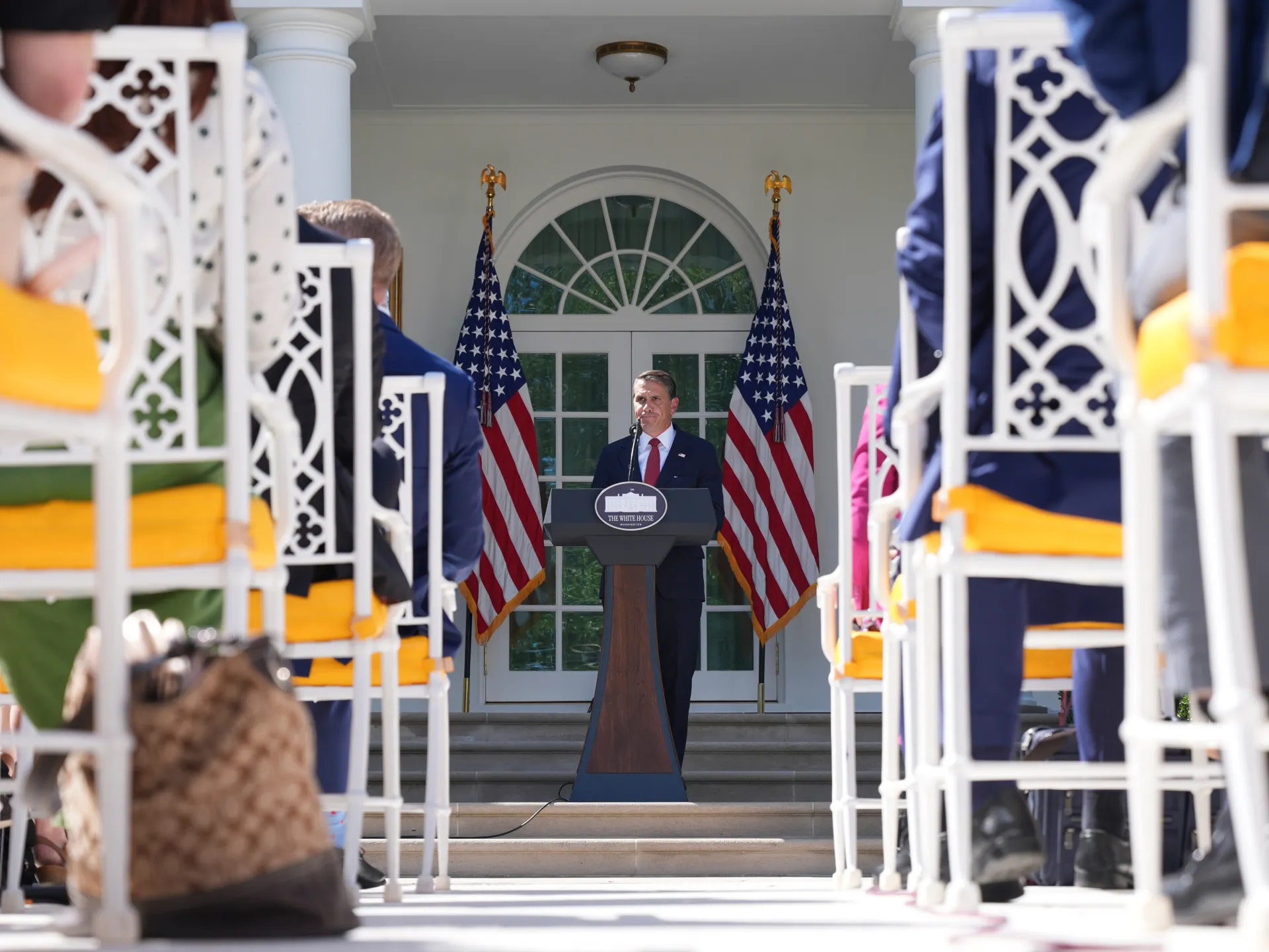 Man speaking at a podium between American flags, viewed through seated audience members