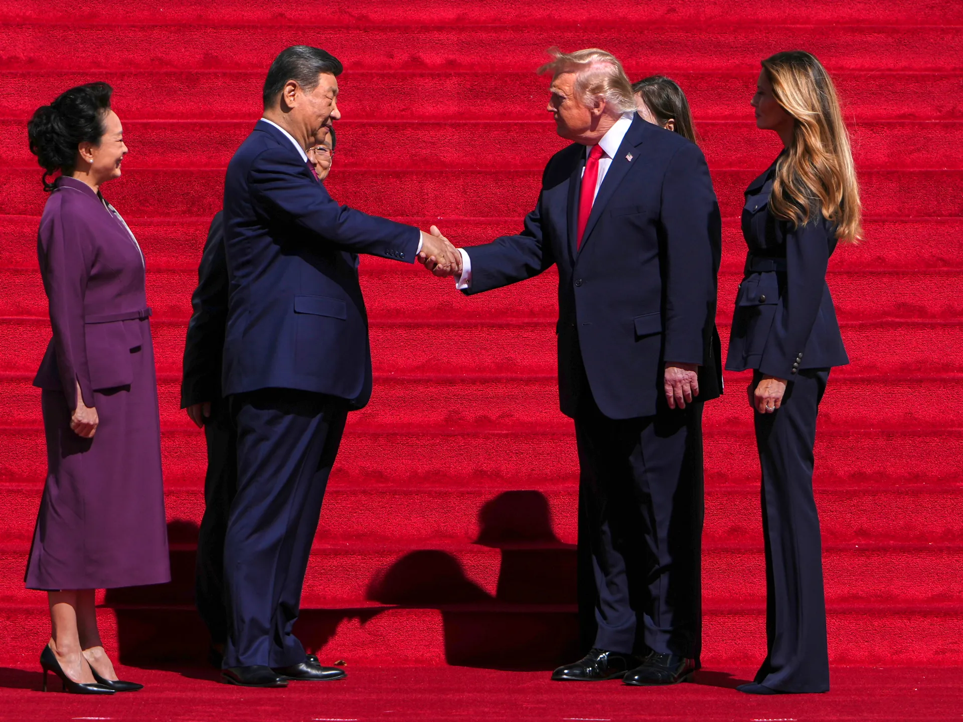 Two men shake hands on red-carpeted steps as three women stand nearby.