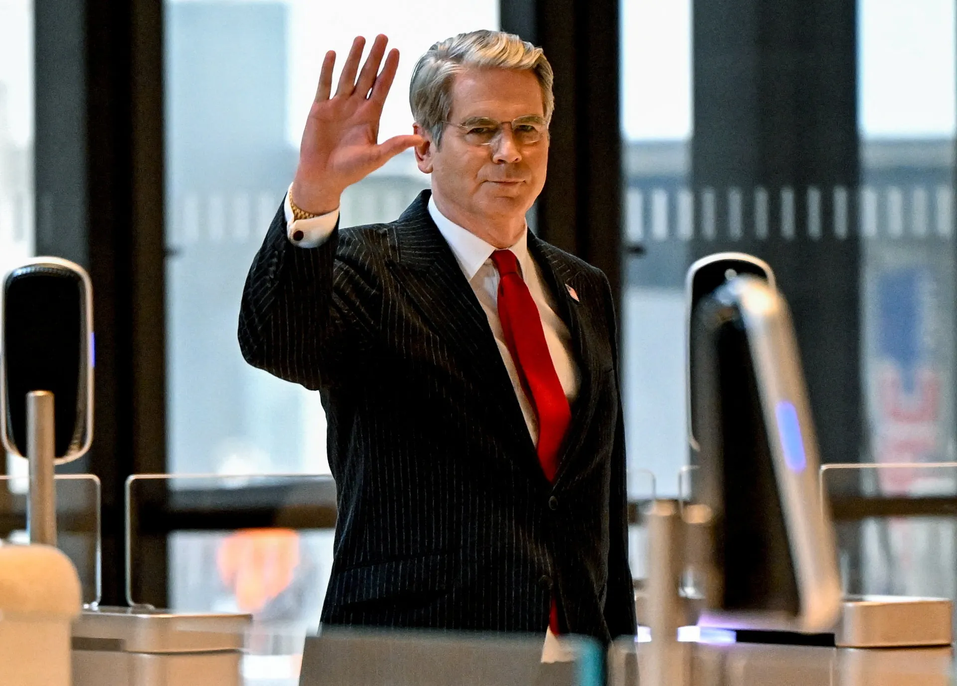 Gray-haired man in a dark pinstripe suit waves near automated security gates.