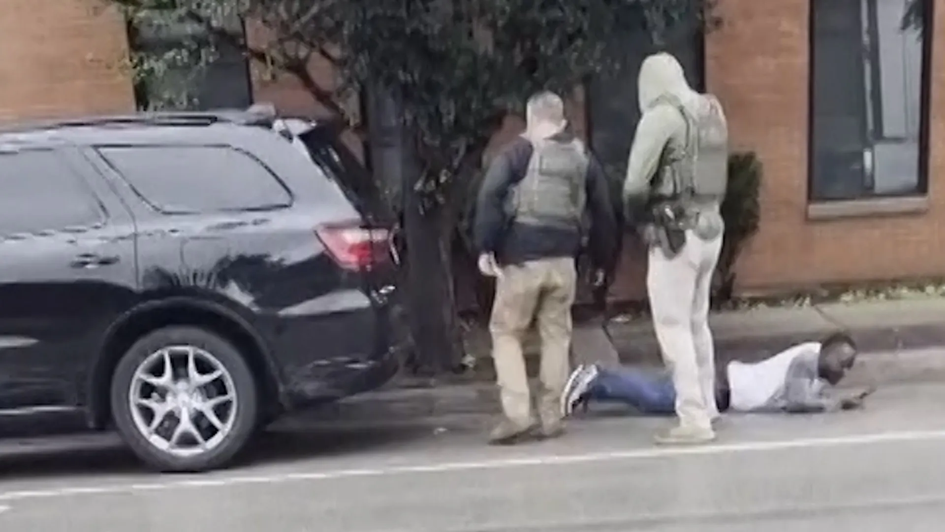 Two people stand beside a dark SUV while another person lies on the pavement near a building.