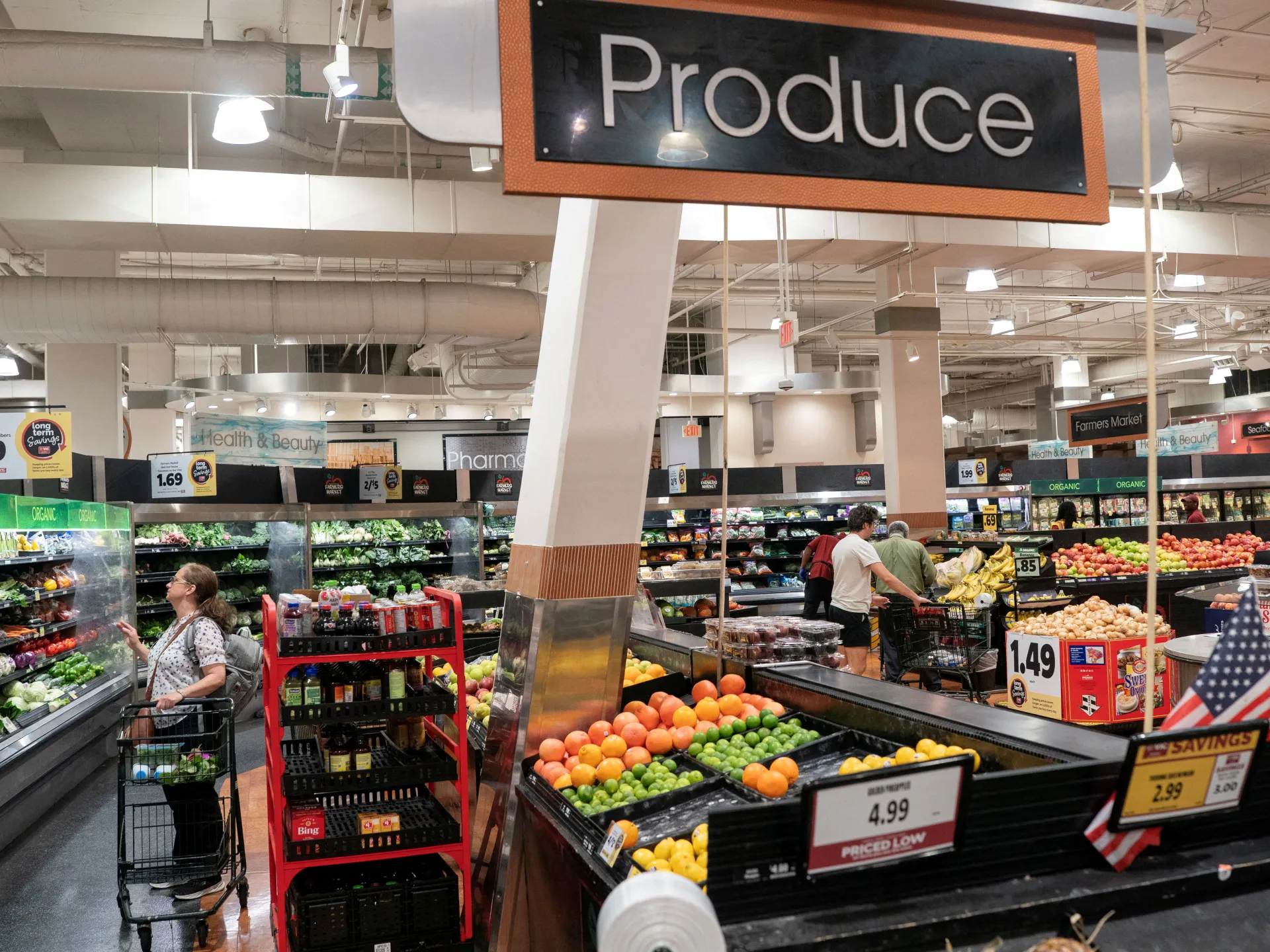 Shoppers browse fresh produce beneath a large sign in a brightly lit grocery store.