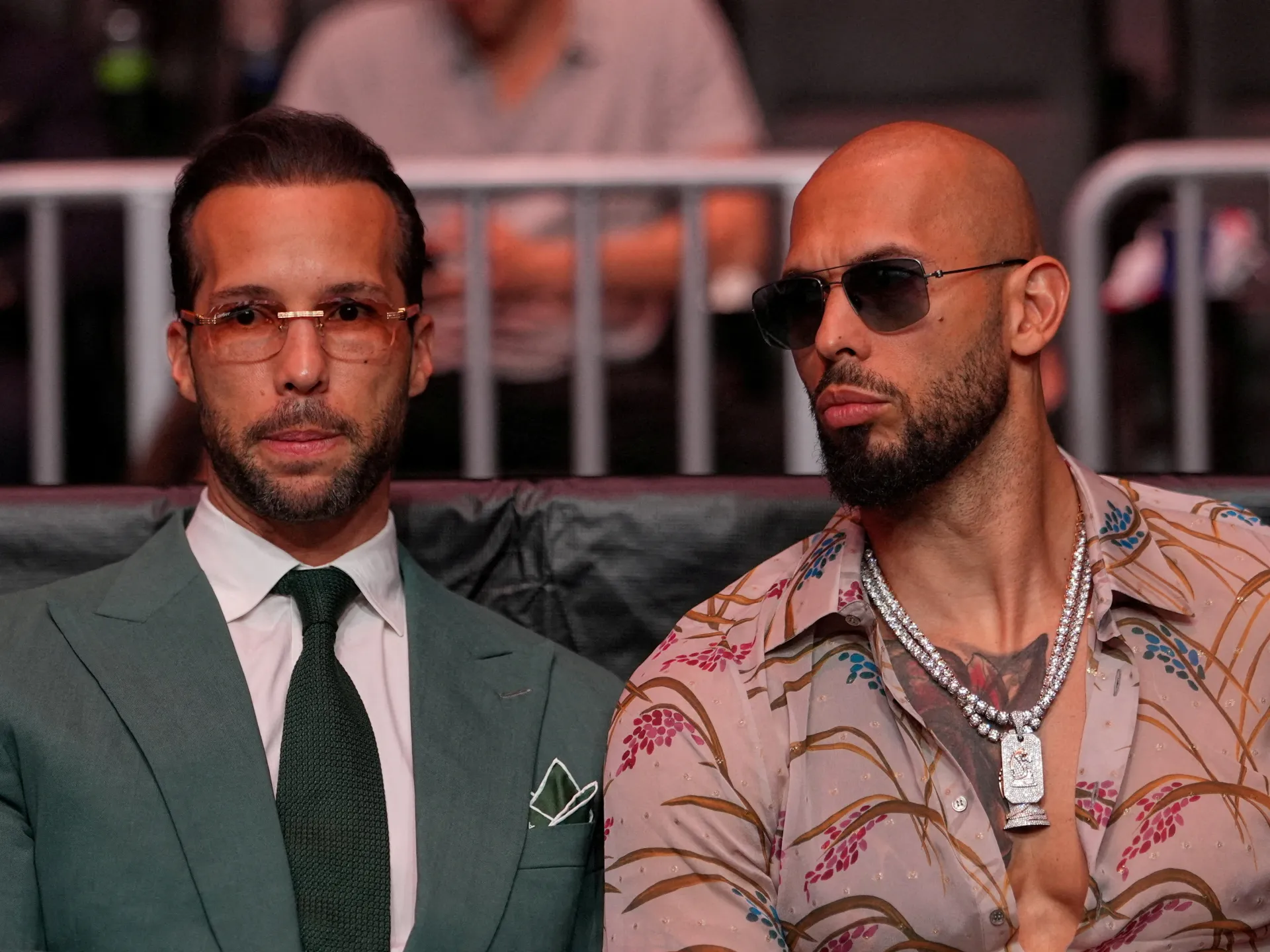 Two men sit side by side, one wearing a suit and the other sunglasses and a patterned shirt.