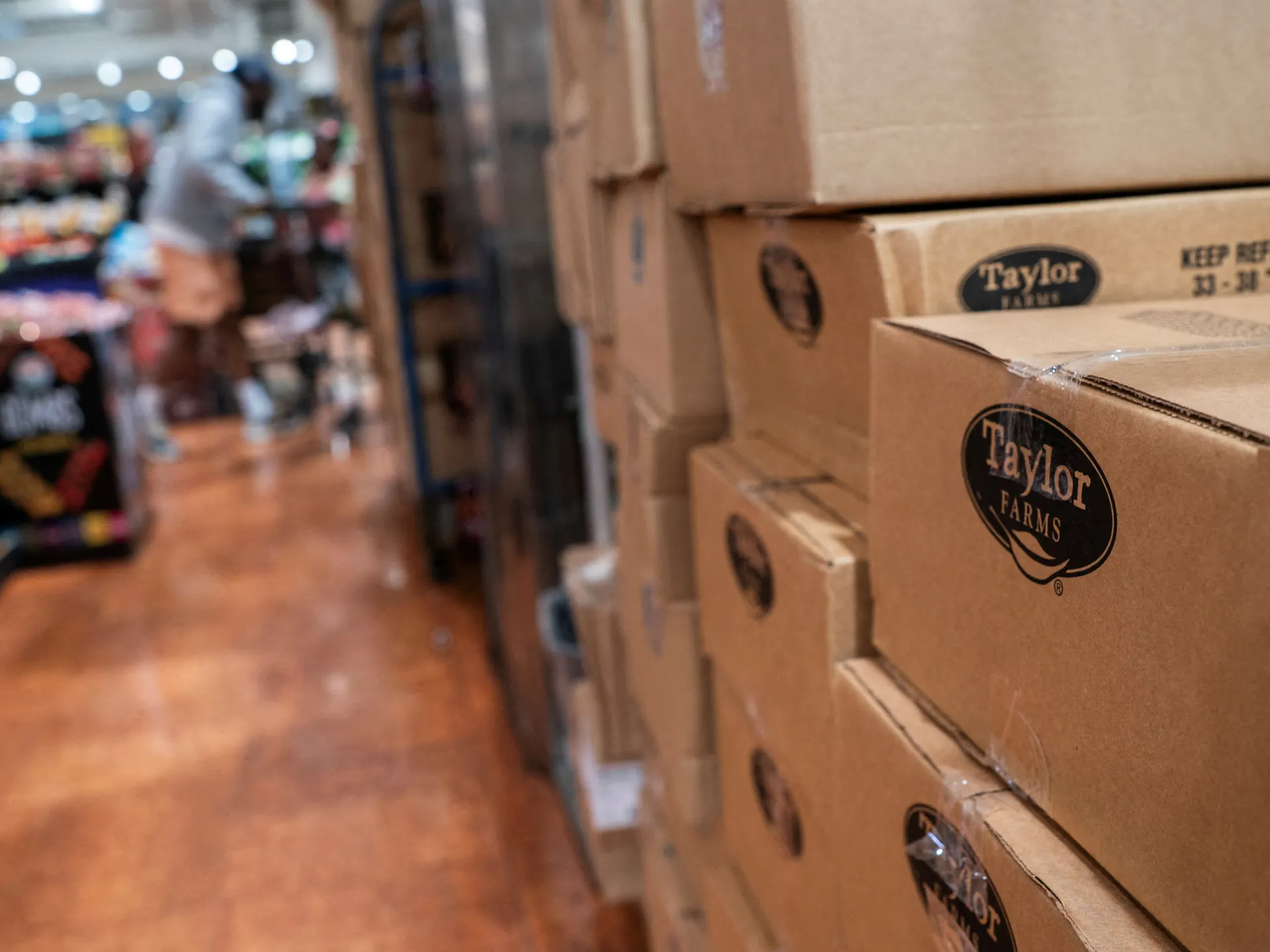 Stacked Taylor Farms cardboard boxes line a warehouse aisle beside blurred workers and produce.