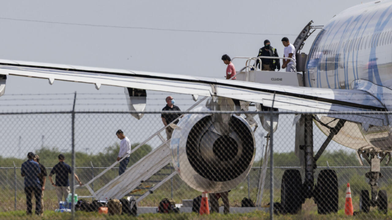 People board and disembark from an airplane using mobile stairs beside a chain-link fence.