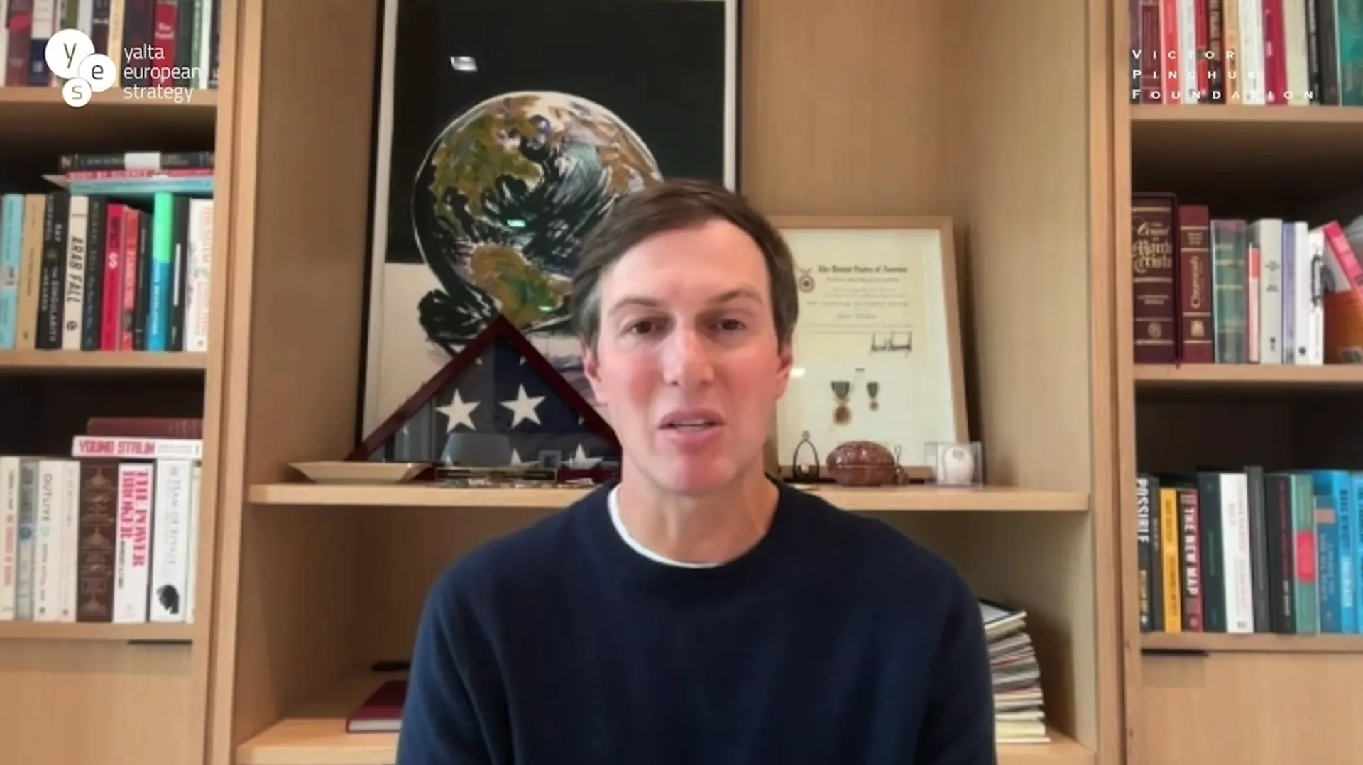Man speaking to camera in front of bookshelves, framed awards, folded flag, and globe artwork.
