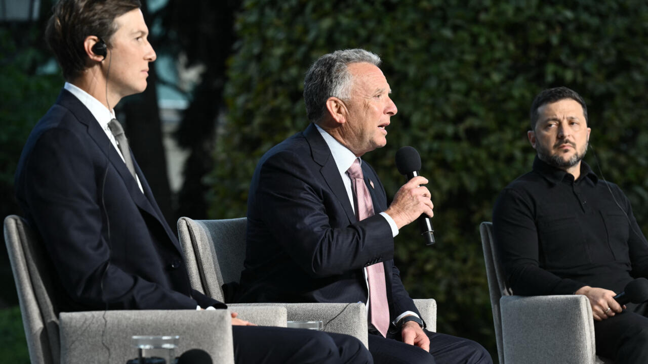Three men sit outdoors during a discussion, with one speaking into a handheld microphone.