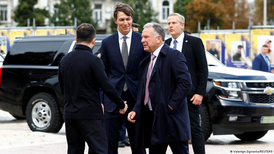 Several men in dark suits and coats stand beside black SUVs, greeting one another outdoors.