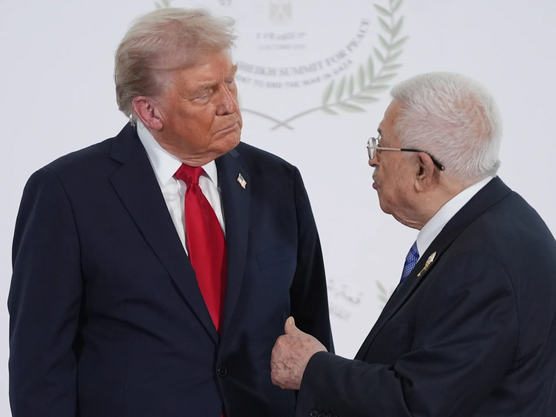 Two older men in dark suits converse, one wearing a red tie and the other glasses.