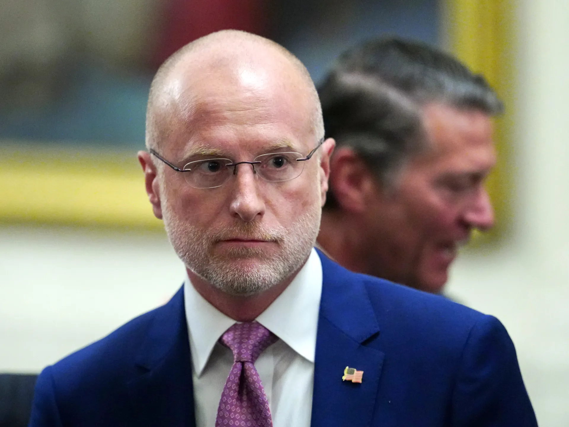 Bald man in glasses and a blue suit stands before a blurred man in a yellow and white room.