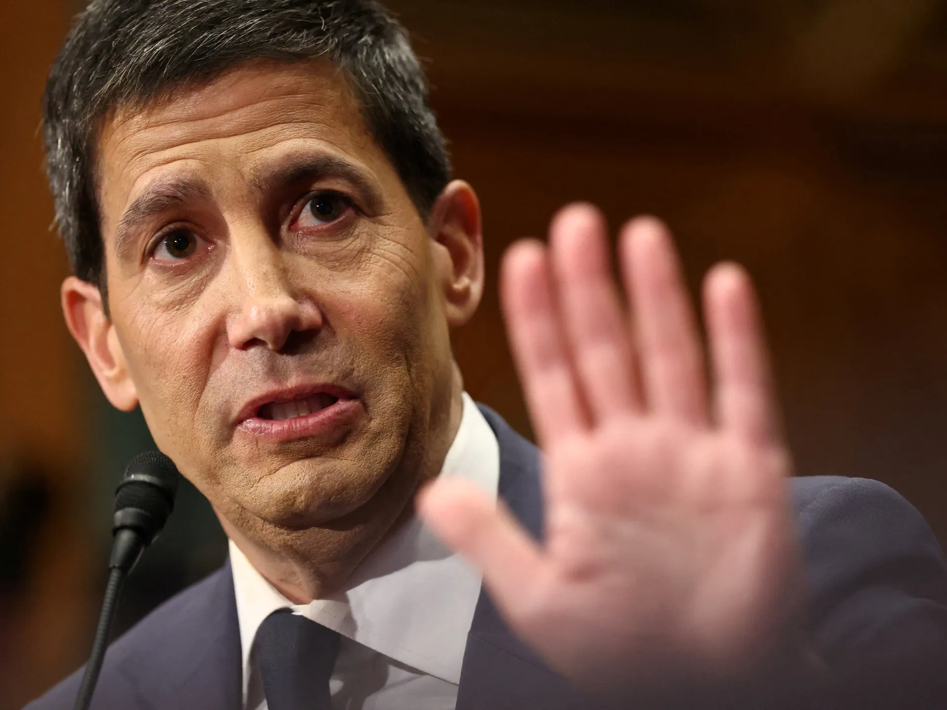 Suited man speaking into a microphone with one hand raised in the foreground