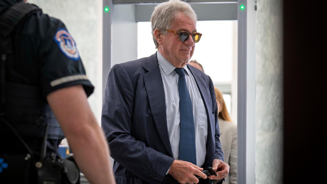 Older man in sunglasses and suit walks past a uniformed security officer indoors