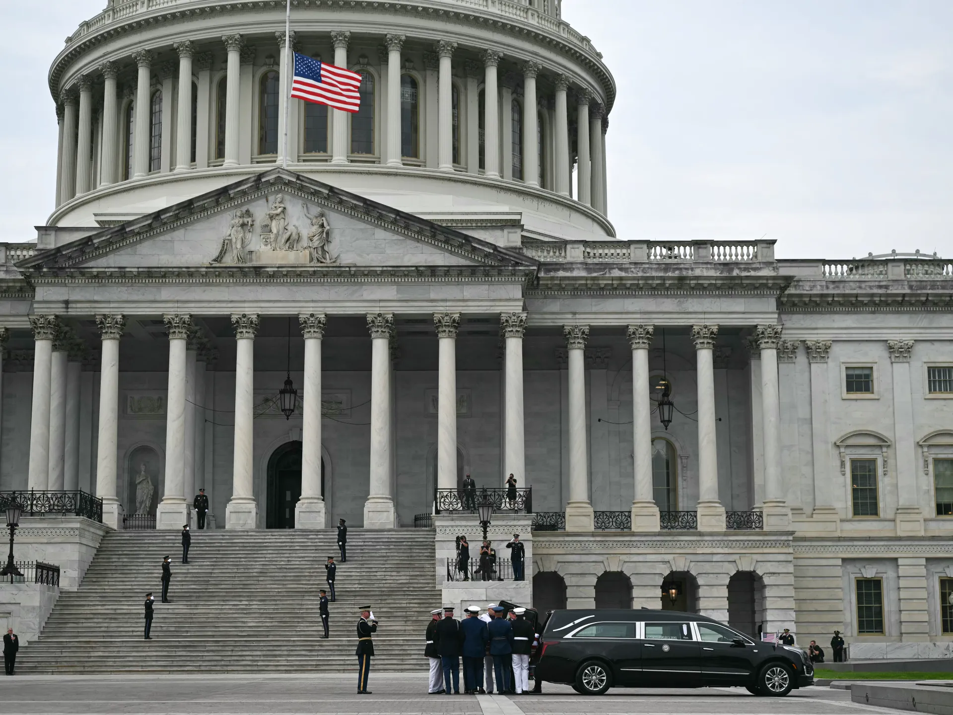 U.S. Capitol portico with lowered American flag, uniformed personnel, and a black hearse outside.