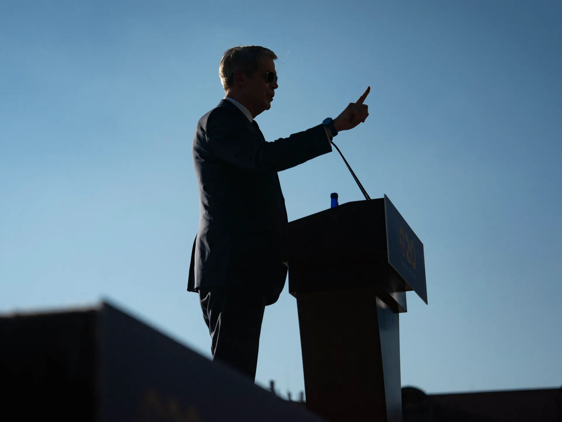Silhouetted suited man gestures beside a podium beneath a clear blue sky.