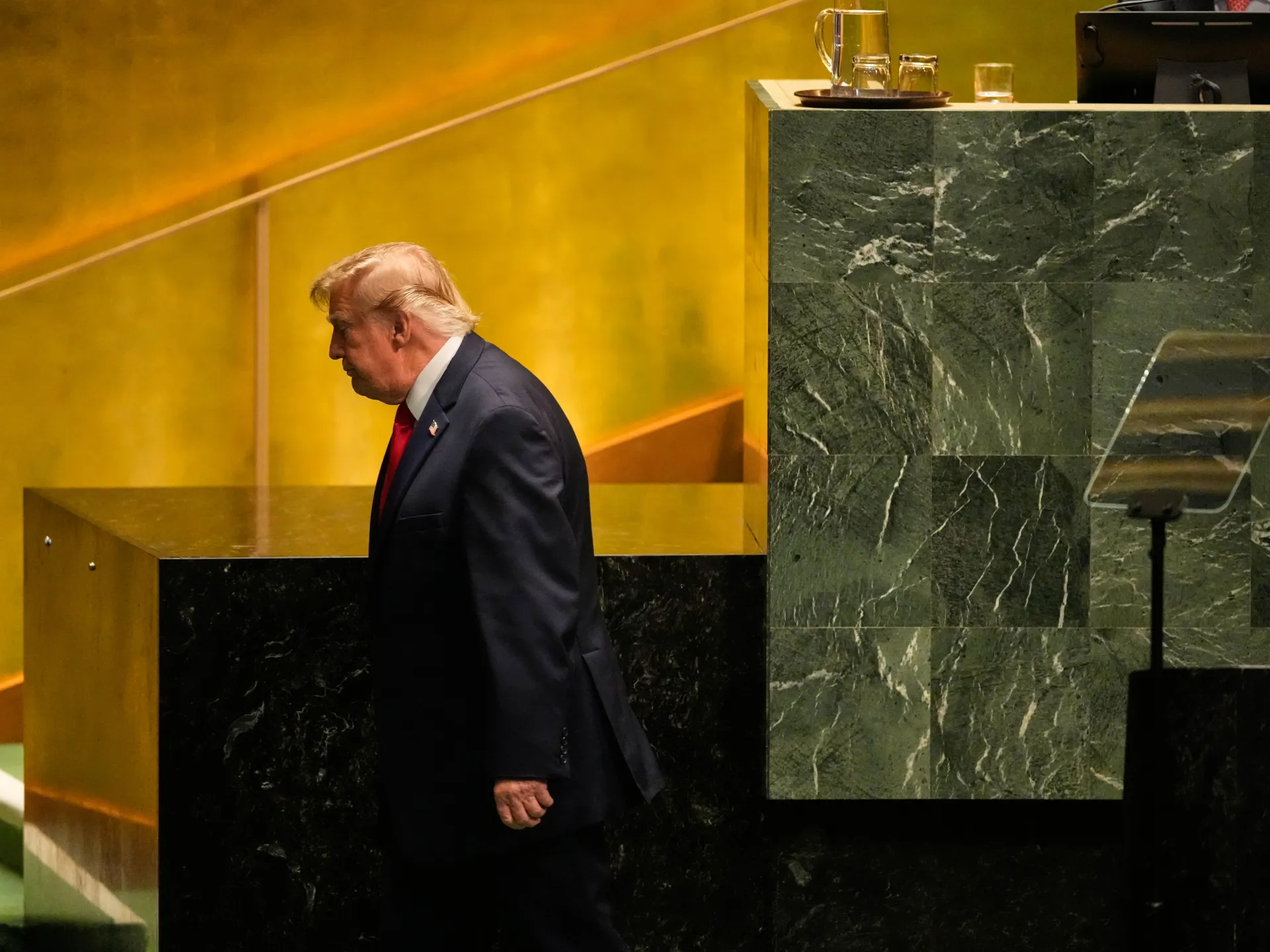An older, blond-haired man in a dark suit stands beside a marble counter in a warmly lit room.