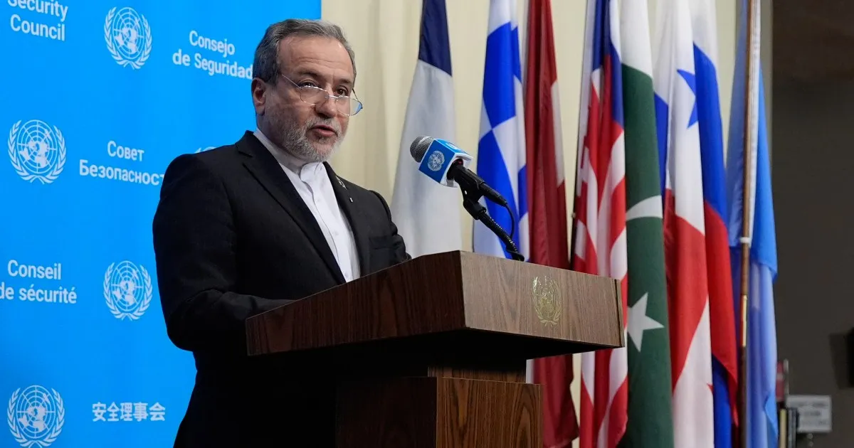 A bearded man in a dark suit speaks at a wooden lectern before United Nations flags.
