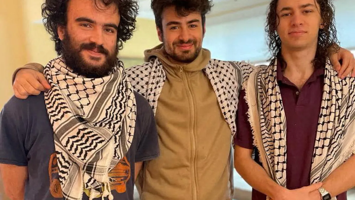 Three men pose indoors, two wearing black-and-white keffiyeh scarves over casual clothing.