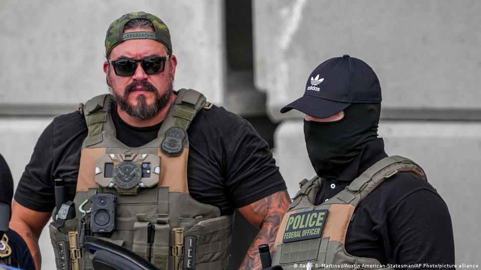 Two armed federal officers in tactical gear stand outside, one wearing a face covering.