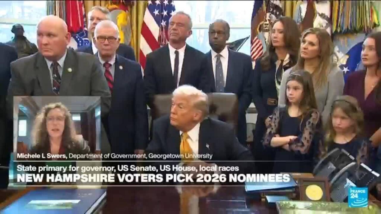 Seated older man faces officials and children during a televised political news broadcast.