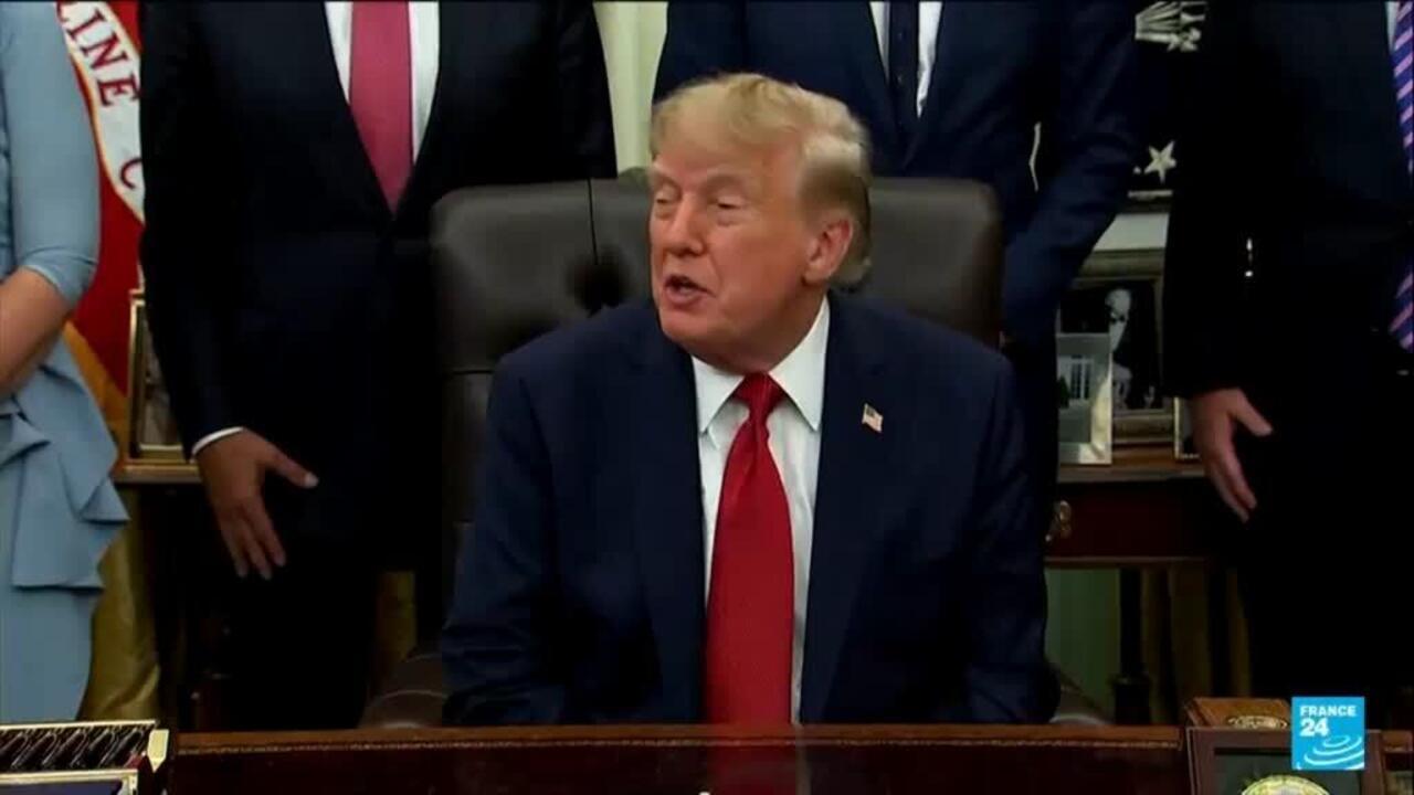 Donald Trump seated at a desk speaking while officials stand behind him