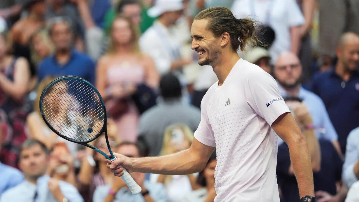 Tennis player with long hair smiles while holding a racket on court before spectators.