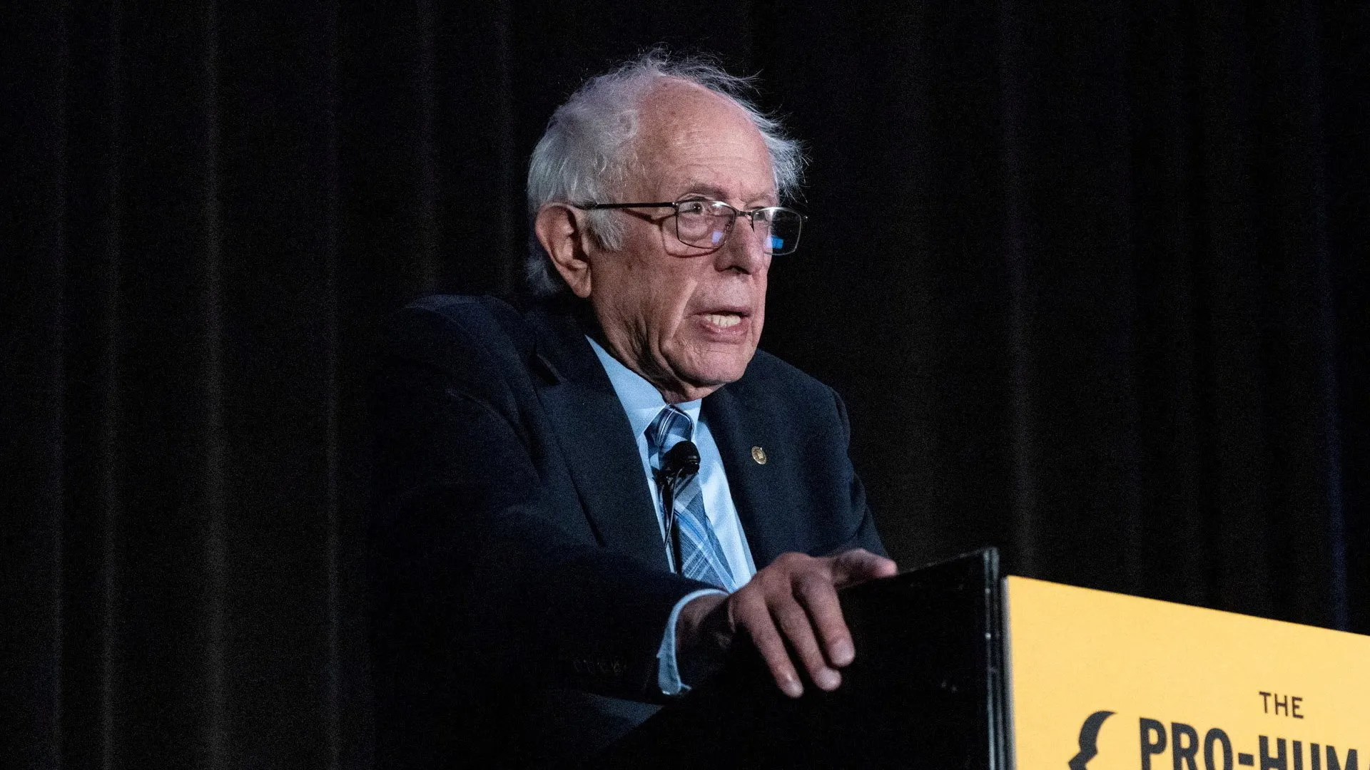 Bernie Sanders speaks at a podium under stage lighting against a dark curtain.