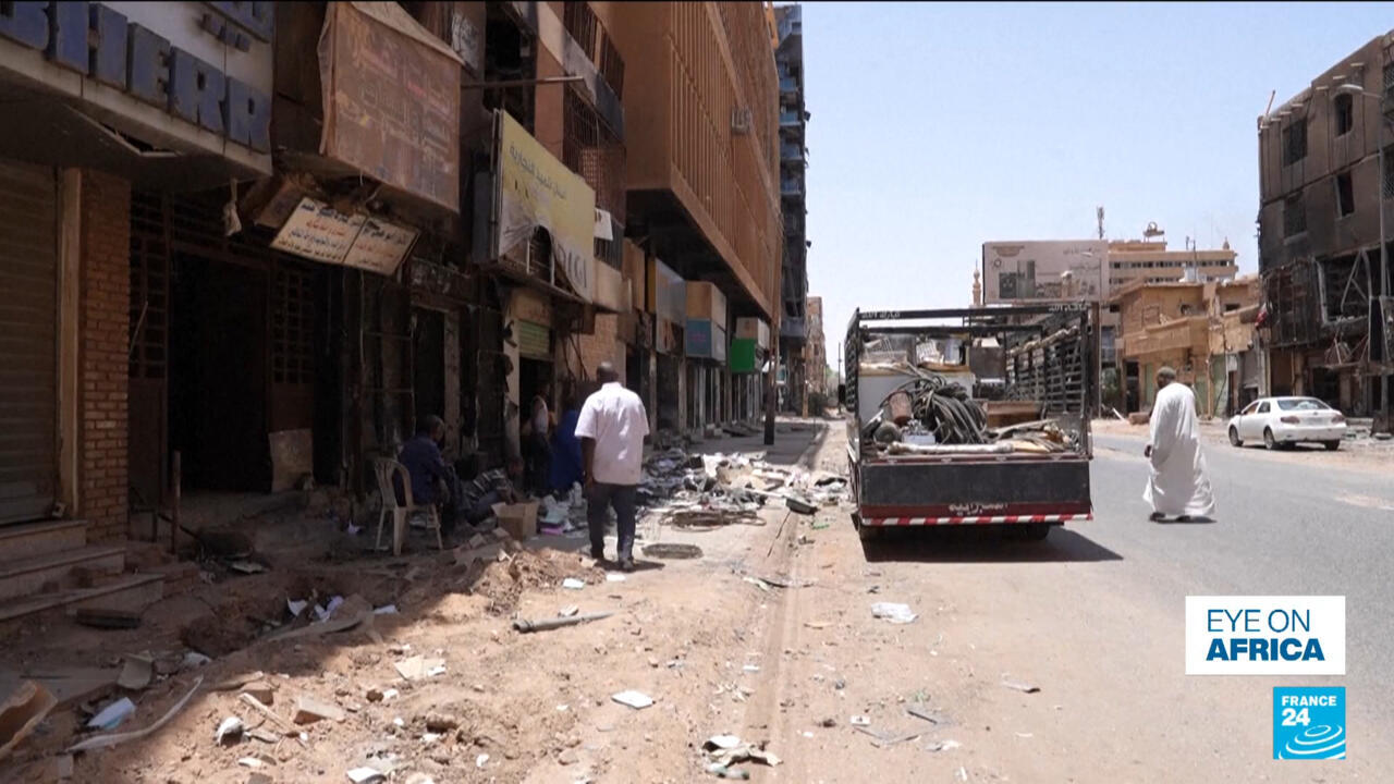 Damaged urban street with debris, pedestrians, storefronts, and a pickup truck carrying scrap materials.