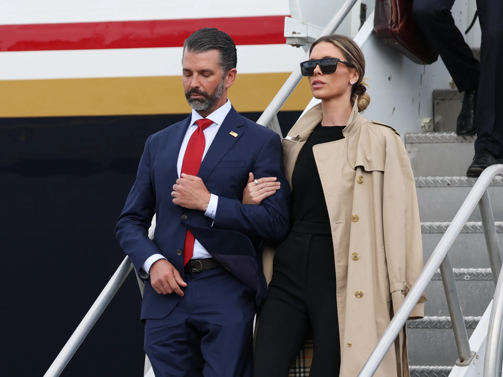 A bearded man in a navy suit and a woman in a beige coat stand on metal stairs.