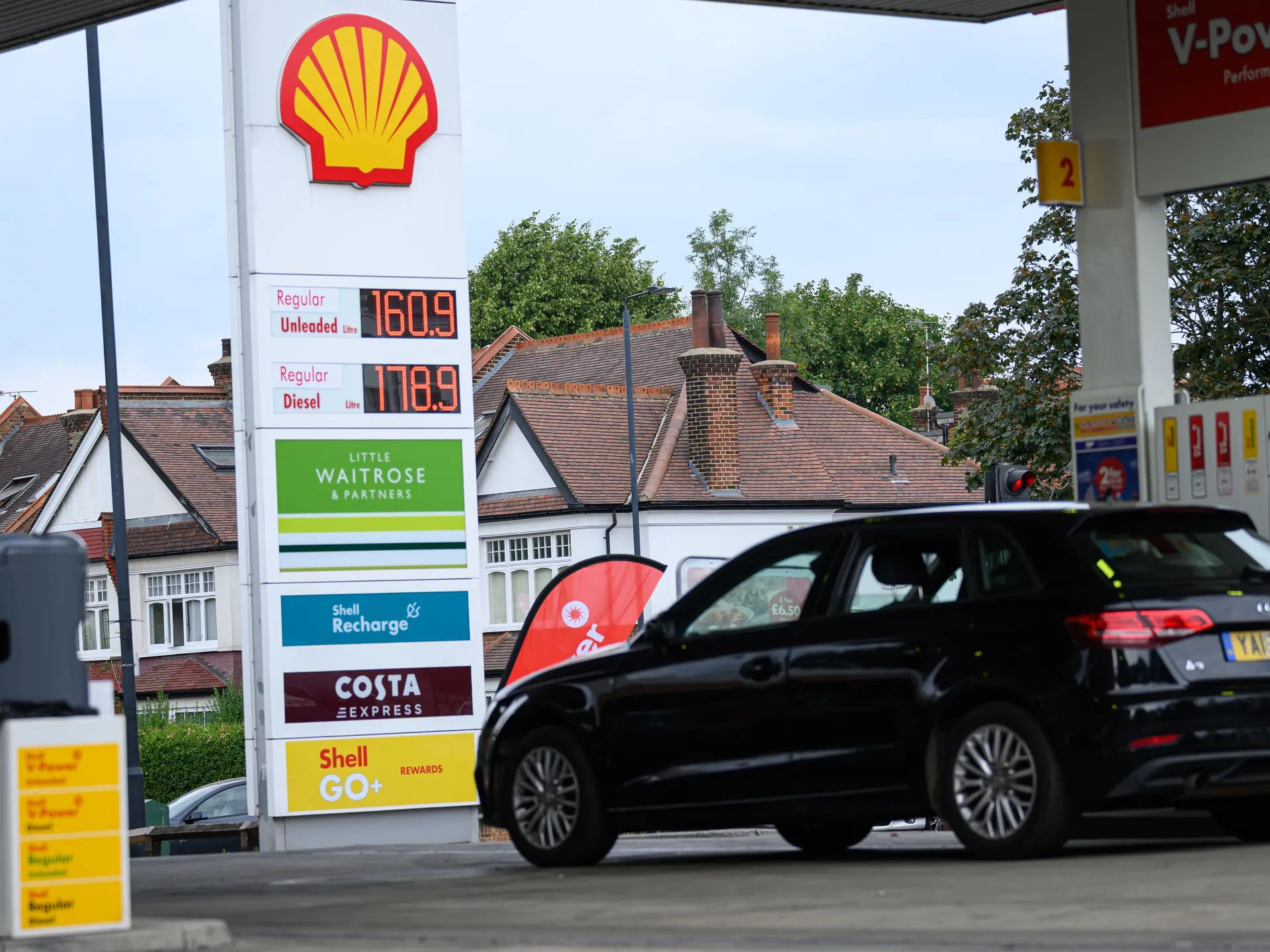 Black car passes a Shell petrol station displaying unleaded and diesel prices beside suburban rooftops.
