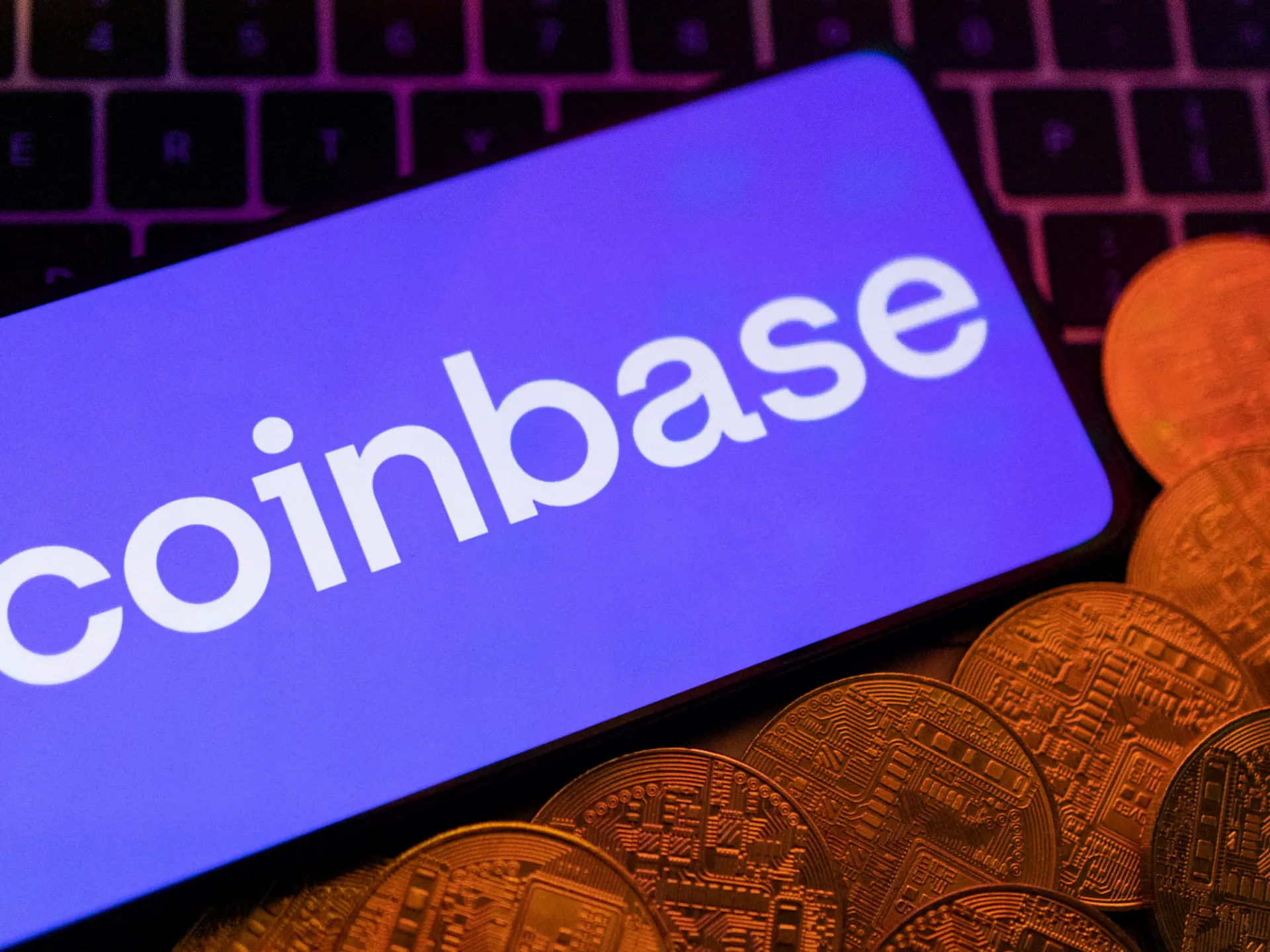 Coinbase logo displayed on a purple device beside cryptocurrency coins and a computer keyboard