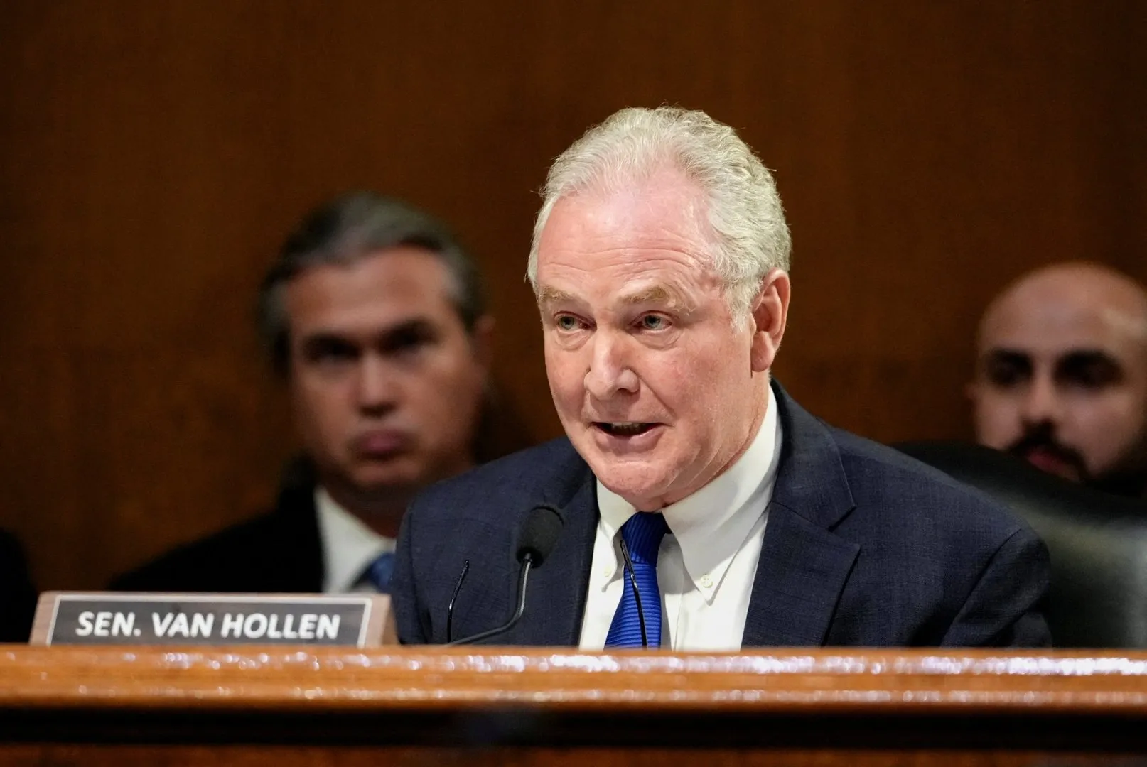 White-haired man in a dark suit speaks at a lectern, with two blurred people behind him.