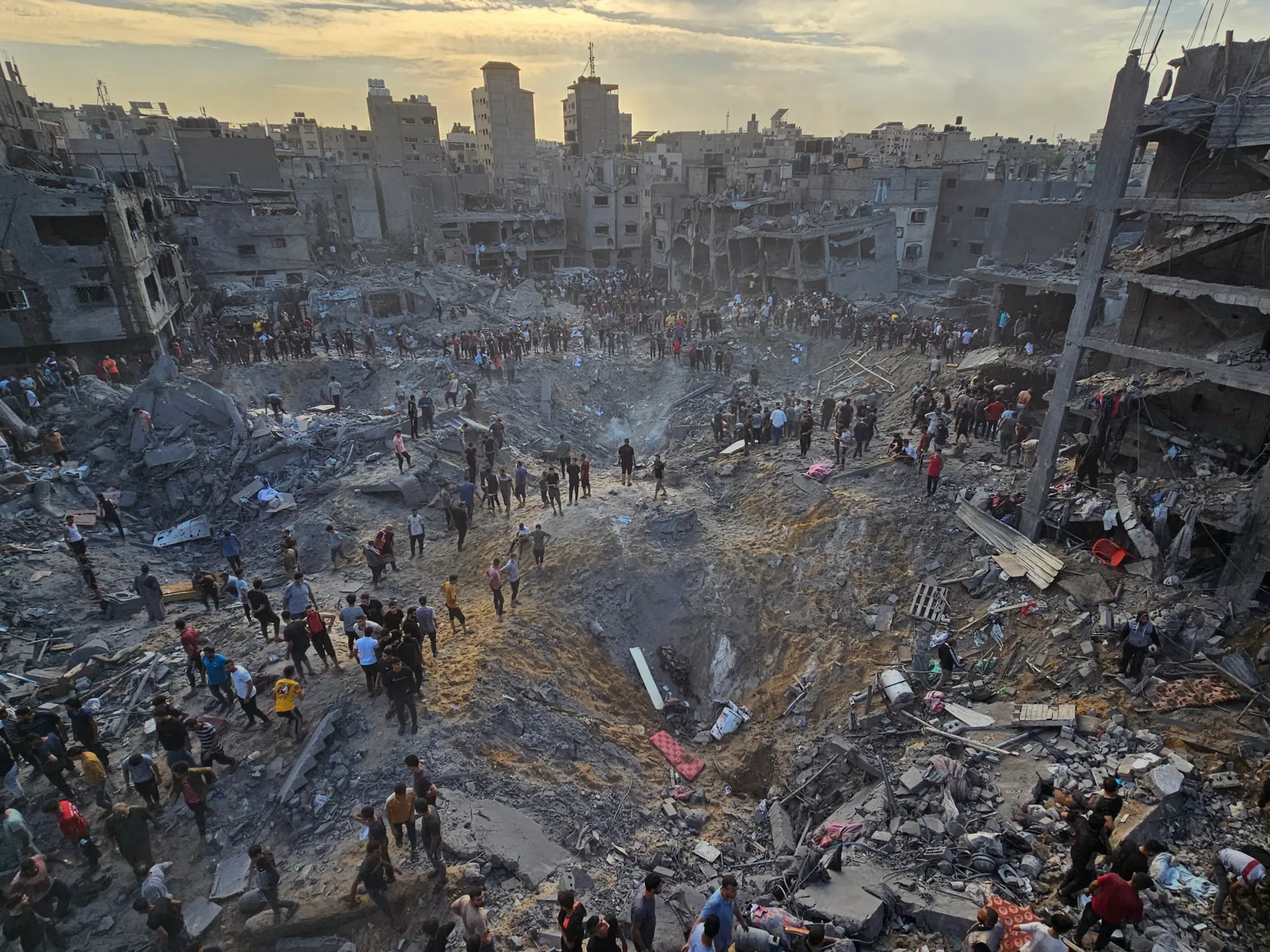 People gather amid extensive rubble and heavily damaged buildings in a densely populated urban area.
