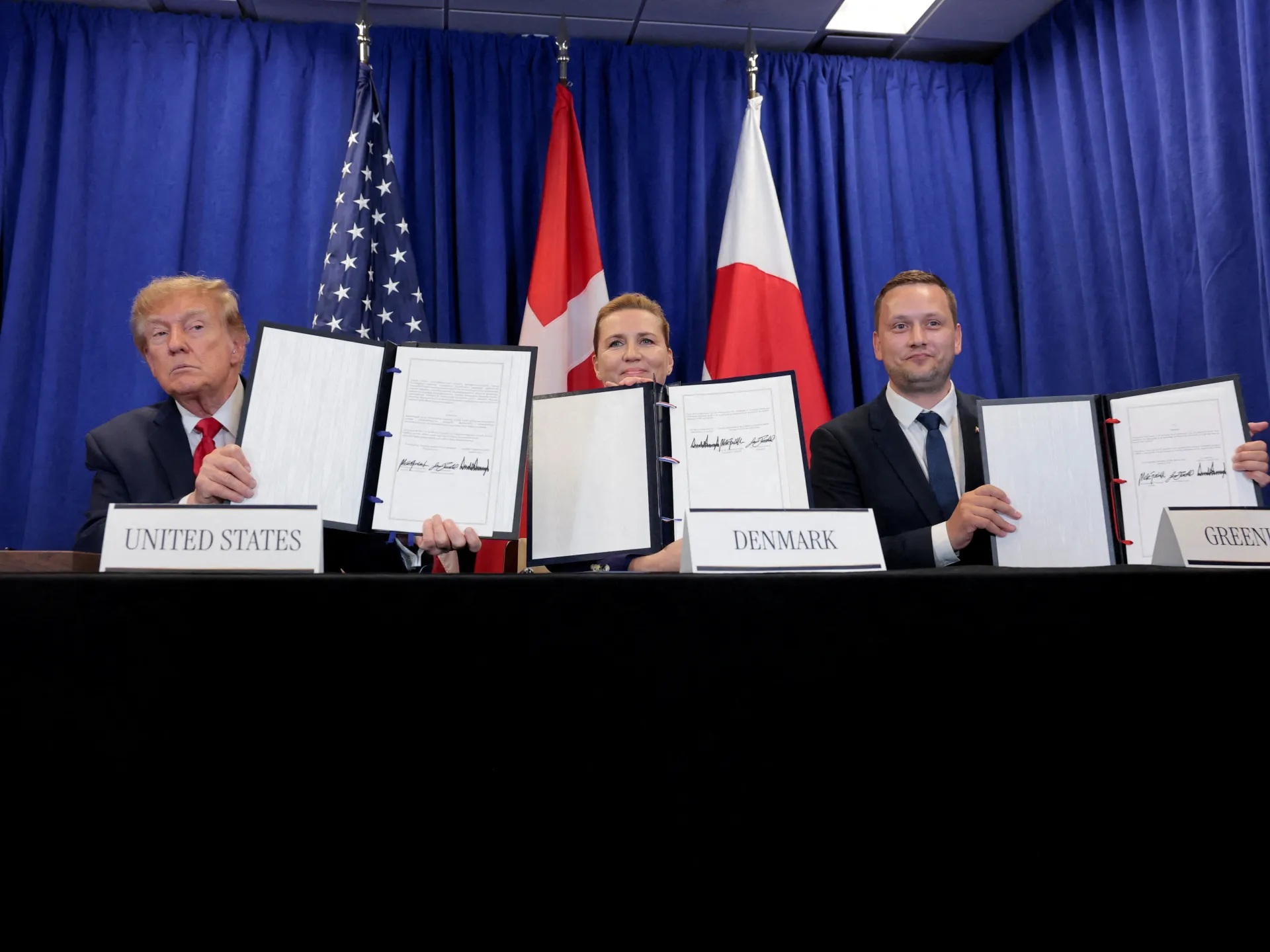 Officials hold signed documents at a table before United States, Danish, and Greenlandic flags.