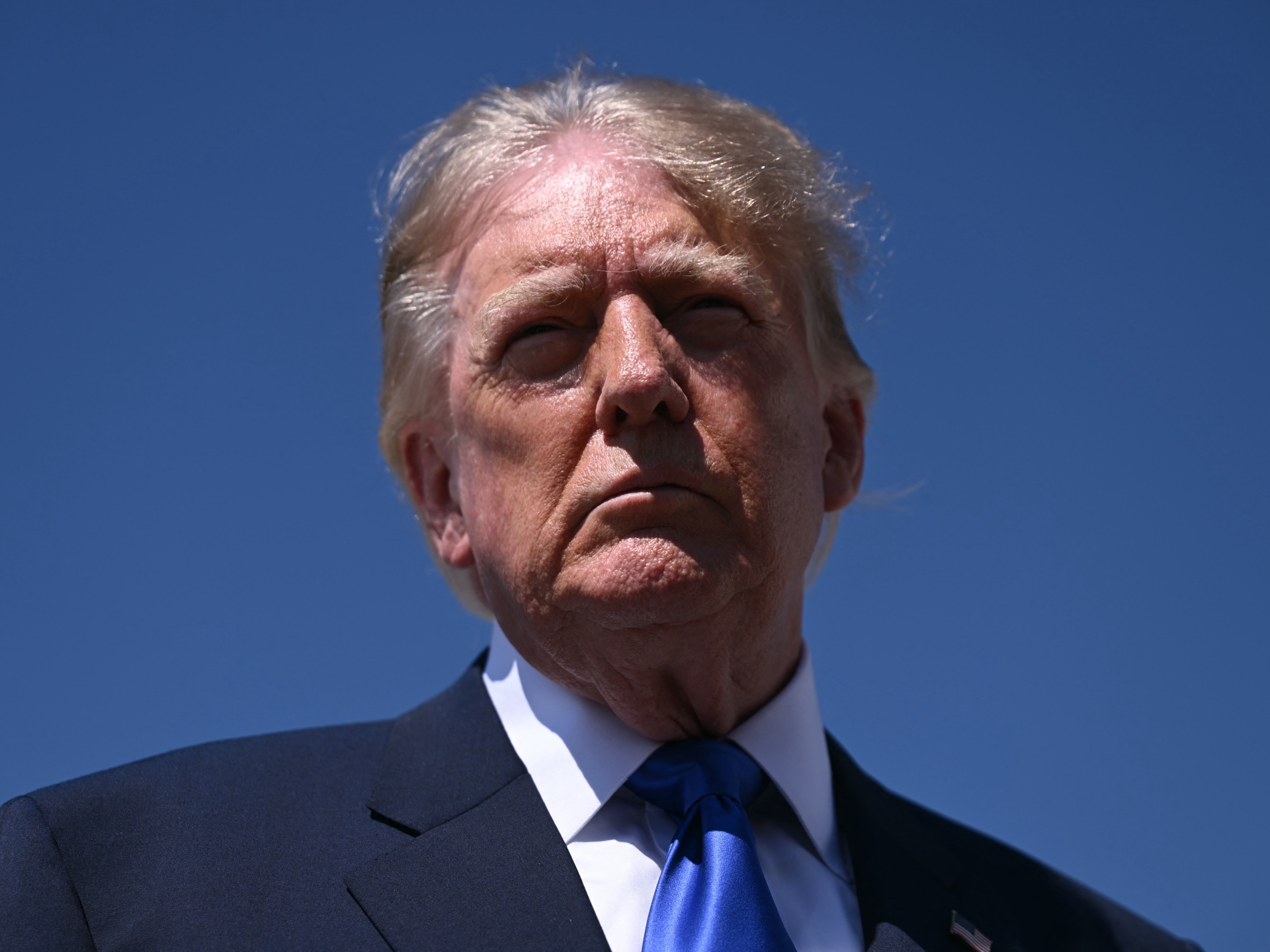 Close-up portrait of Donald Trump in a dark suit and blue tie against a blue sky