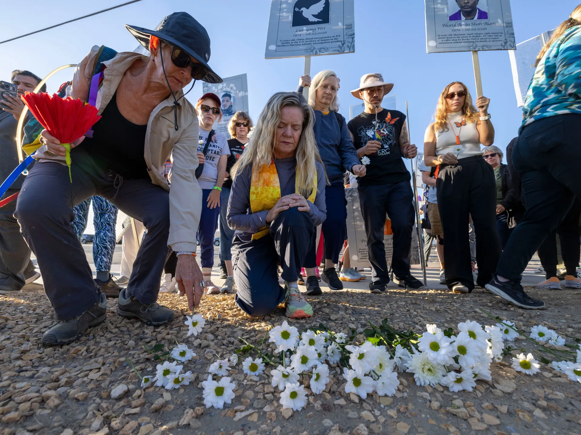 Mourners kneel among white flowers as others hold signs and place flowers on rocky ground.