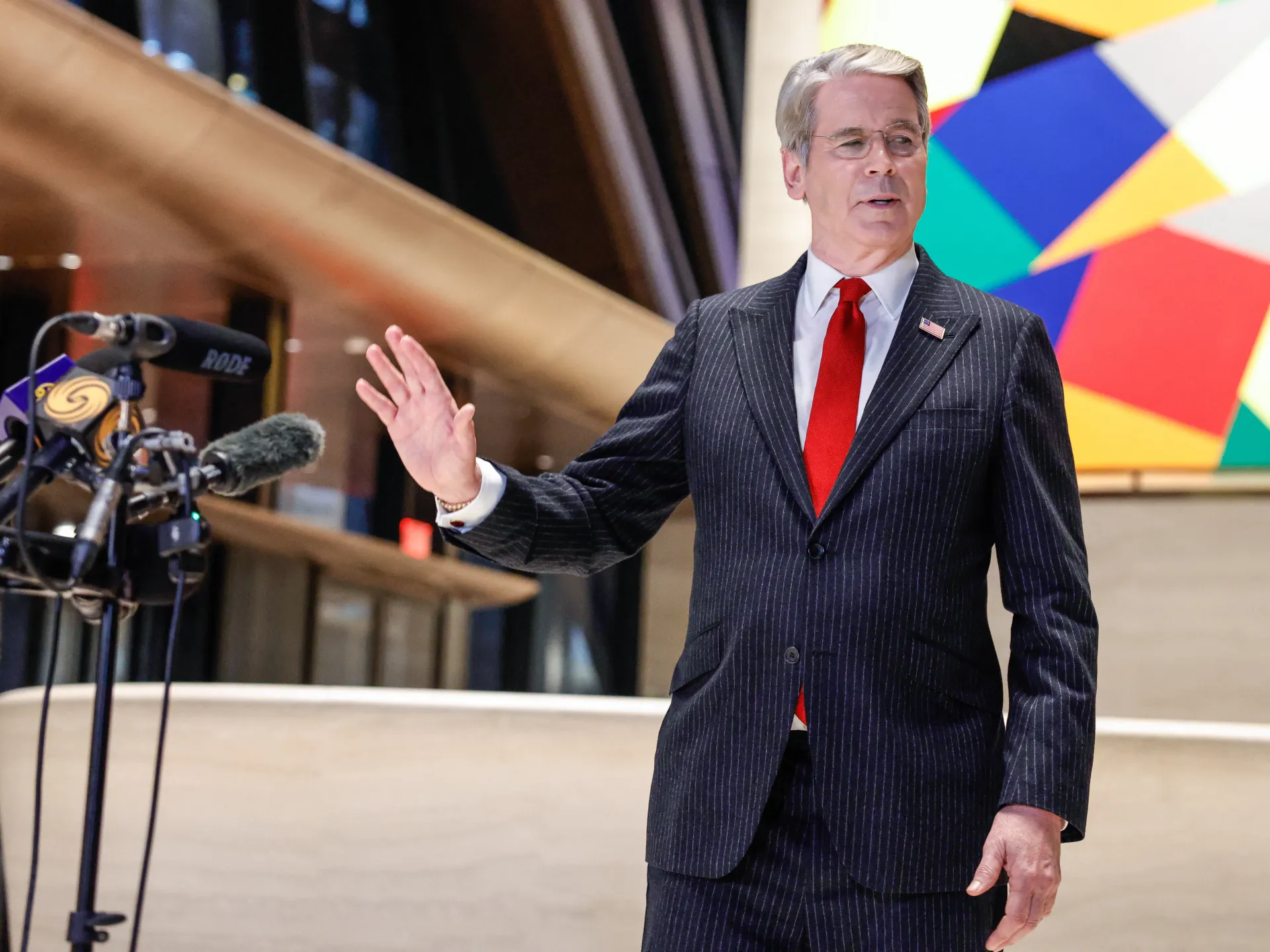 Gray-haired man in pinstripe suit gestures beside microphones in a modern interior