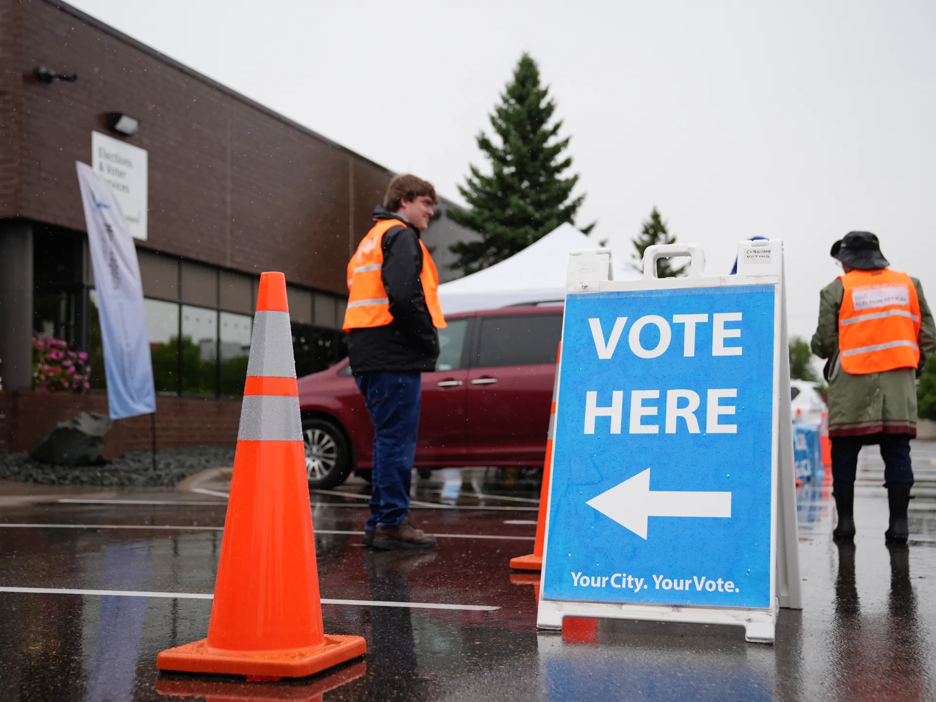 A roadside sign reading “Vote Here” points left beside election workers in reflective vests.