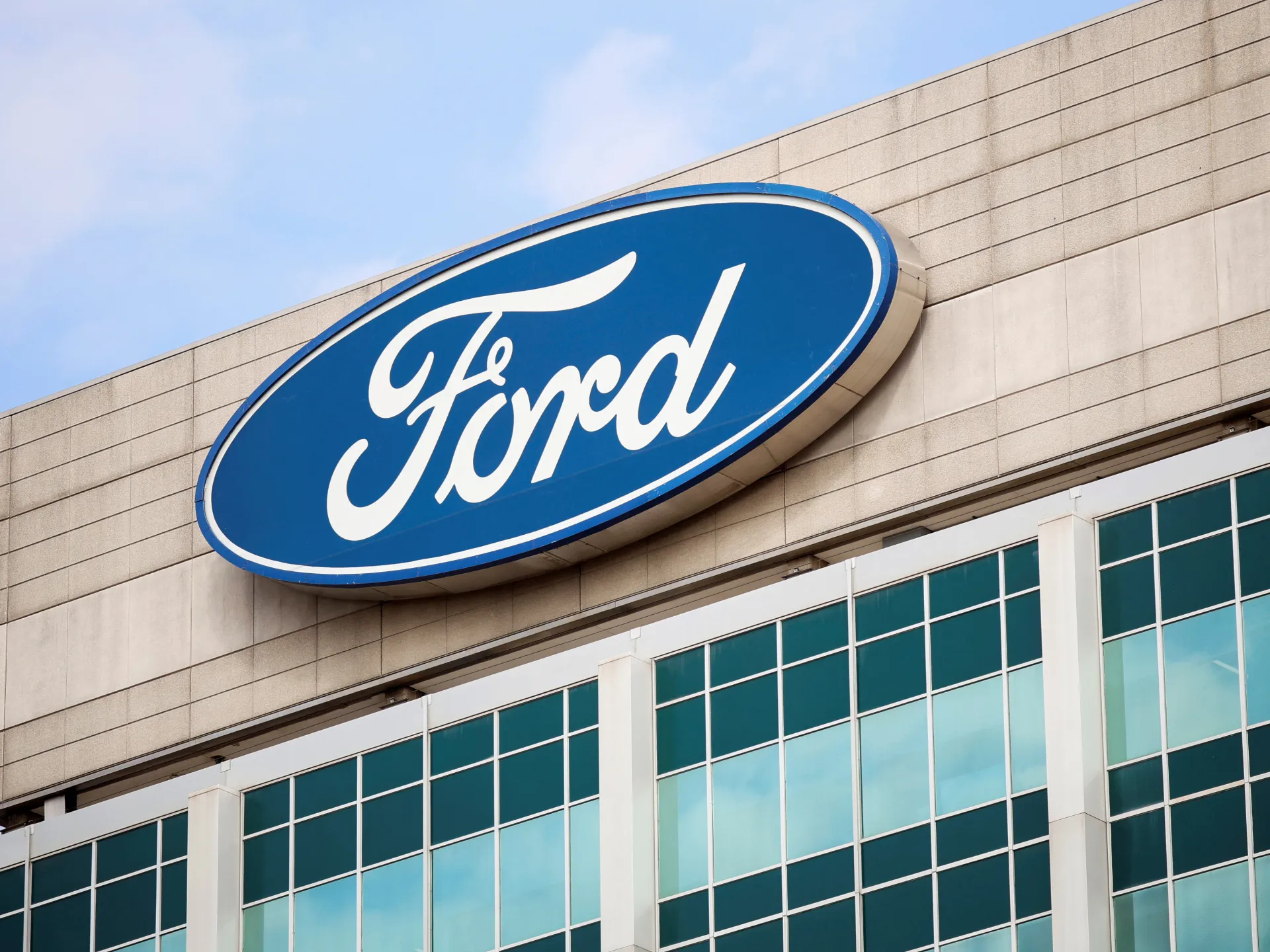 Ford logo mounted on a building facade above large glass windows under a blue sky