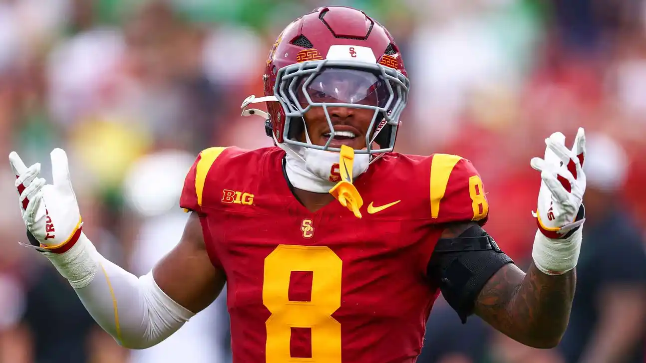 USC football player in a red and gold uniform raises both gloved hands on the field.