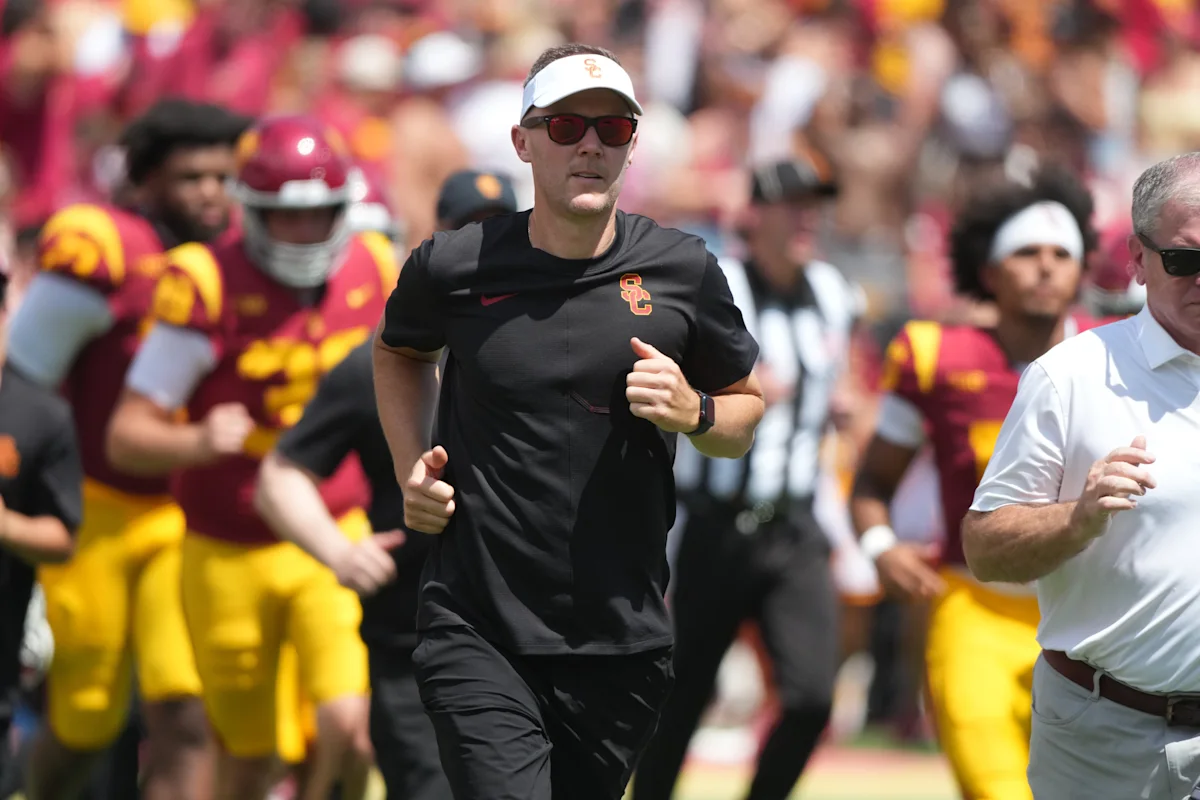USC football coach in black athletic wear runs alongside players during a game