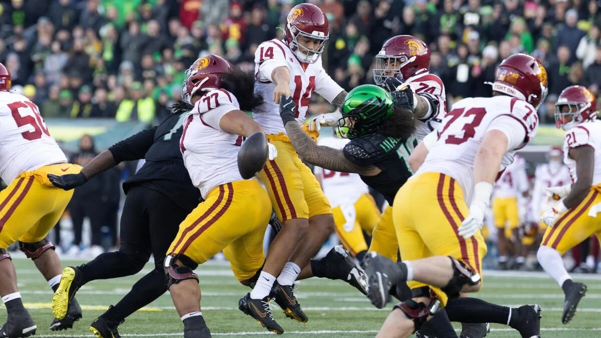 USC players in white and gold block an Oregon player in black and green during a football play.