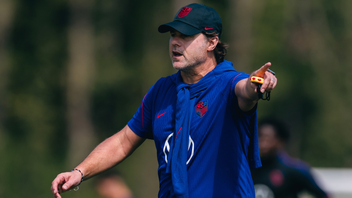 Man in a blue U.S. soccer training shirt and cap points while speaking on a field.