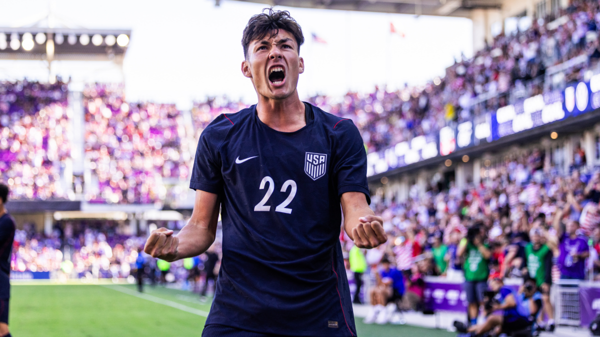 Young soccer player in a navy number 22 jersey celebrates with clenched fists before a packed stadium crowd.