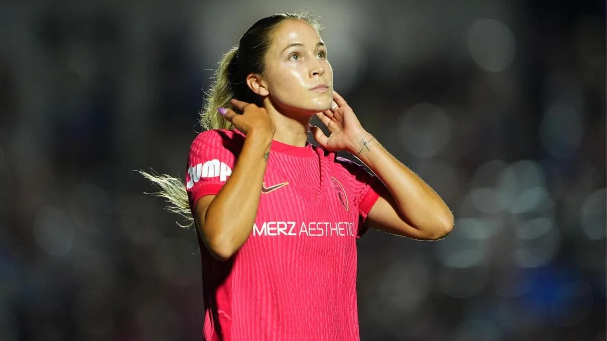 A woman in a bright pink soccer jersey raises both hands near her neck under stadium lights.