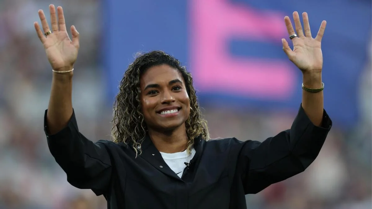 A smiling soccer player raises both hands during an onstage appearance.