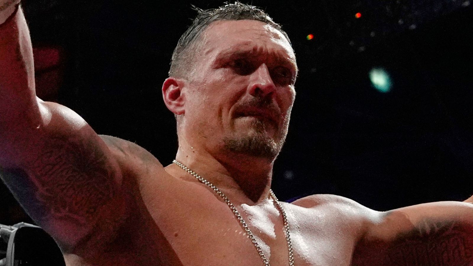 Muscular shirtless male boxer raises both arms under dramatic arena lighting.