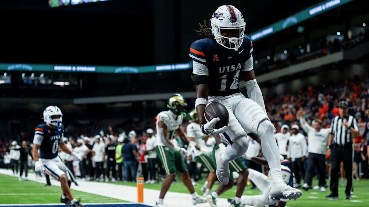 UTSA player in a navy jersey leaps with the football near the sideline as defenders trail.