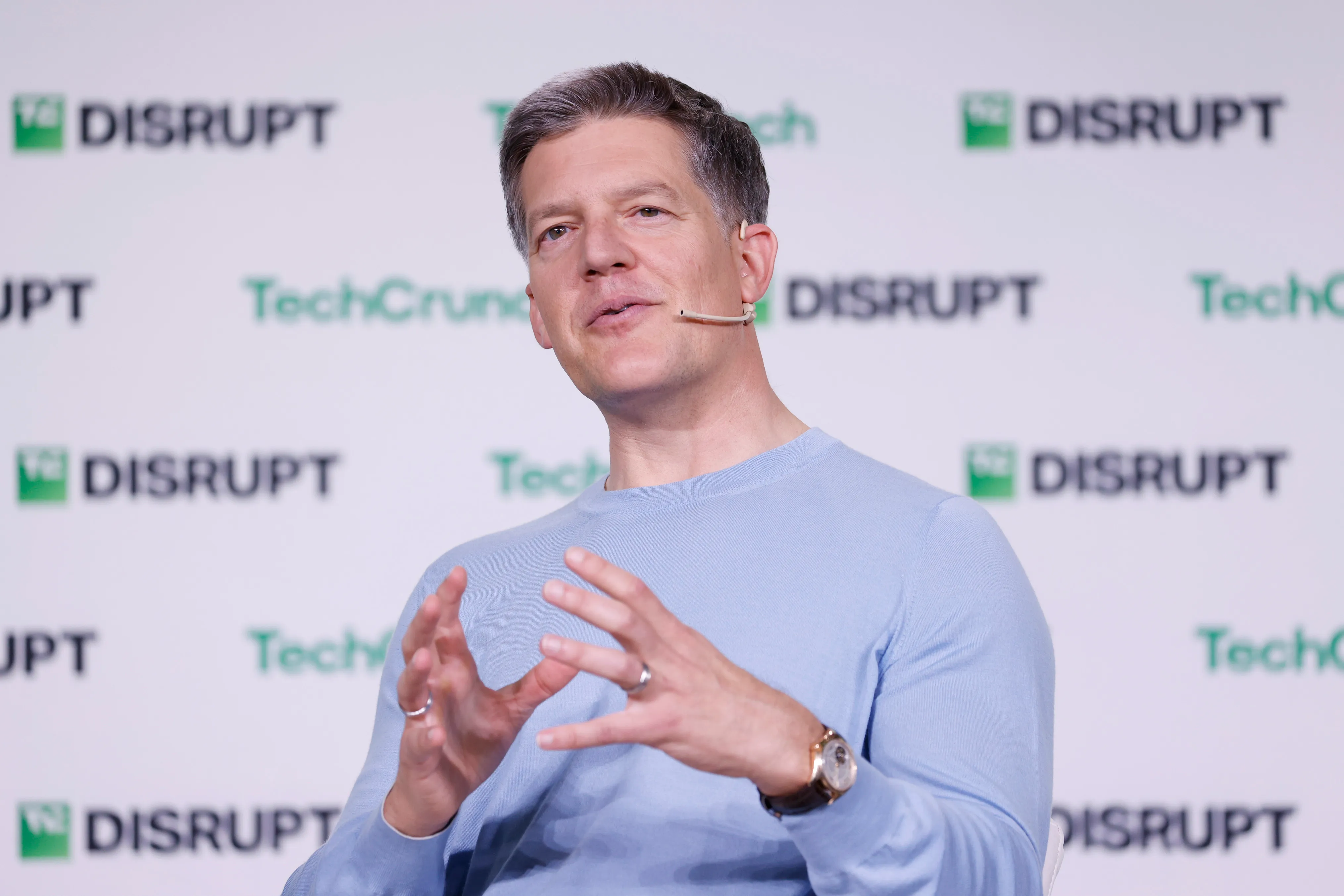 Man in a light blue sweater speaks onstage, gesturing in front of a TechCrunch Disrupt backdrop.