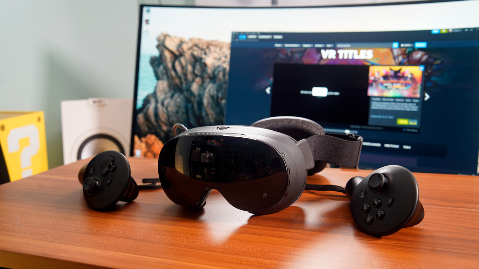 Standalone VR headset and two controllers on a wooden desk before a computer monitor.