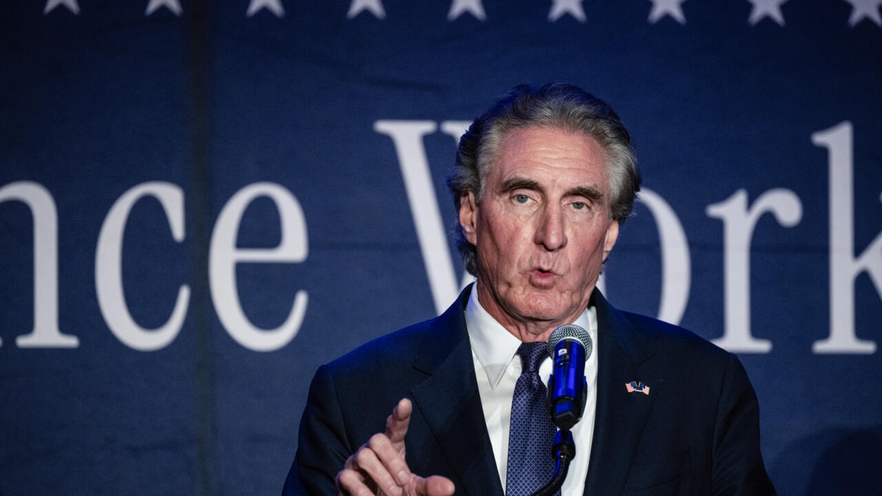 Gray-haired man in suit speaking into a microphone against a dark blue backdrop